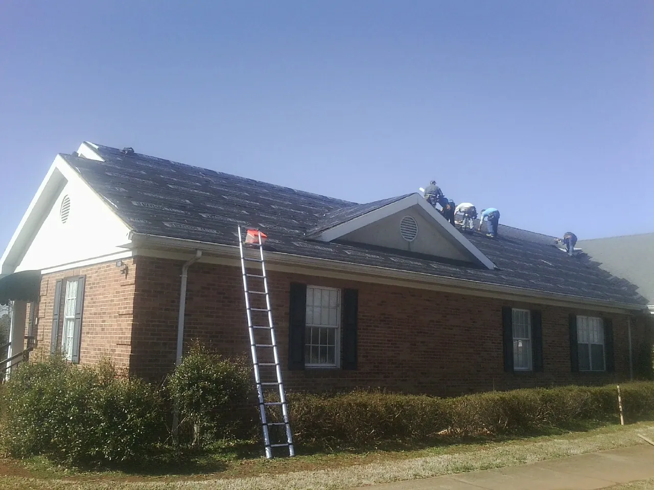 Crew tearing shingles off a brick ranch roof with a ladder set — Emergency Roof Repair work by our crew in Cayuta