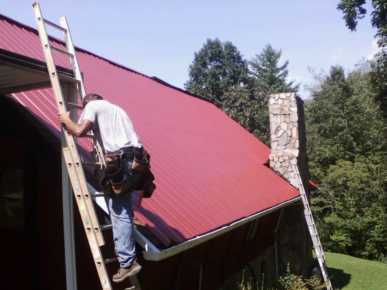 Roofer on a ladder beside a red metal roof and stone chimney — Soffit & Fascia Repair by Roofing Brothers in Cayuta